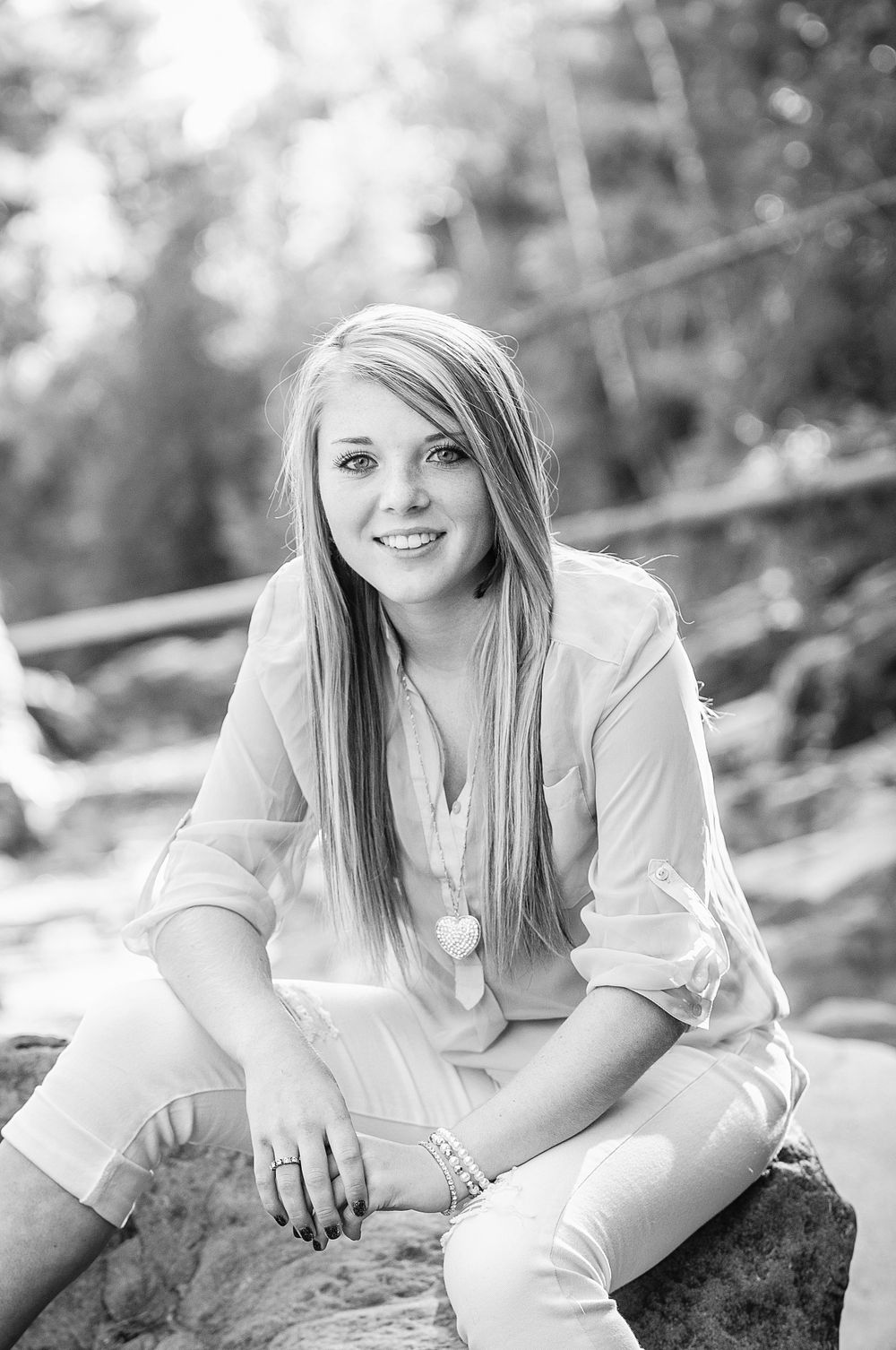 Black and white senior portrait of a smiling blonde girl sitting on rocks wearing a light blouse and white jeans.