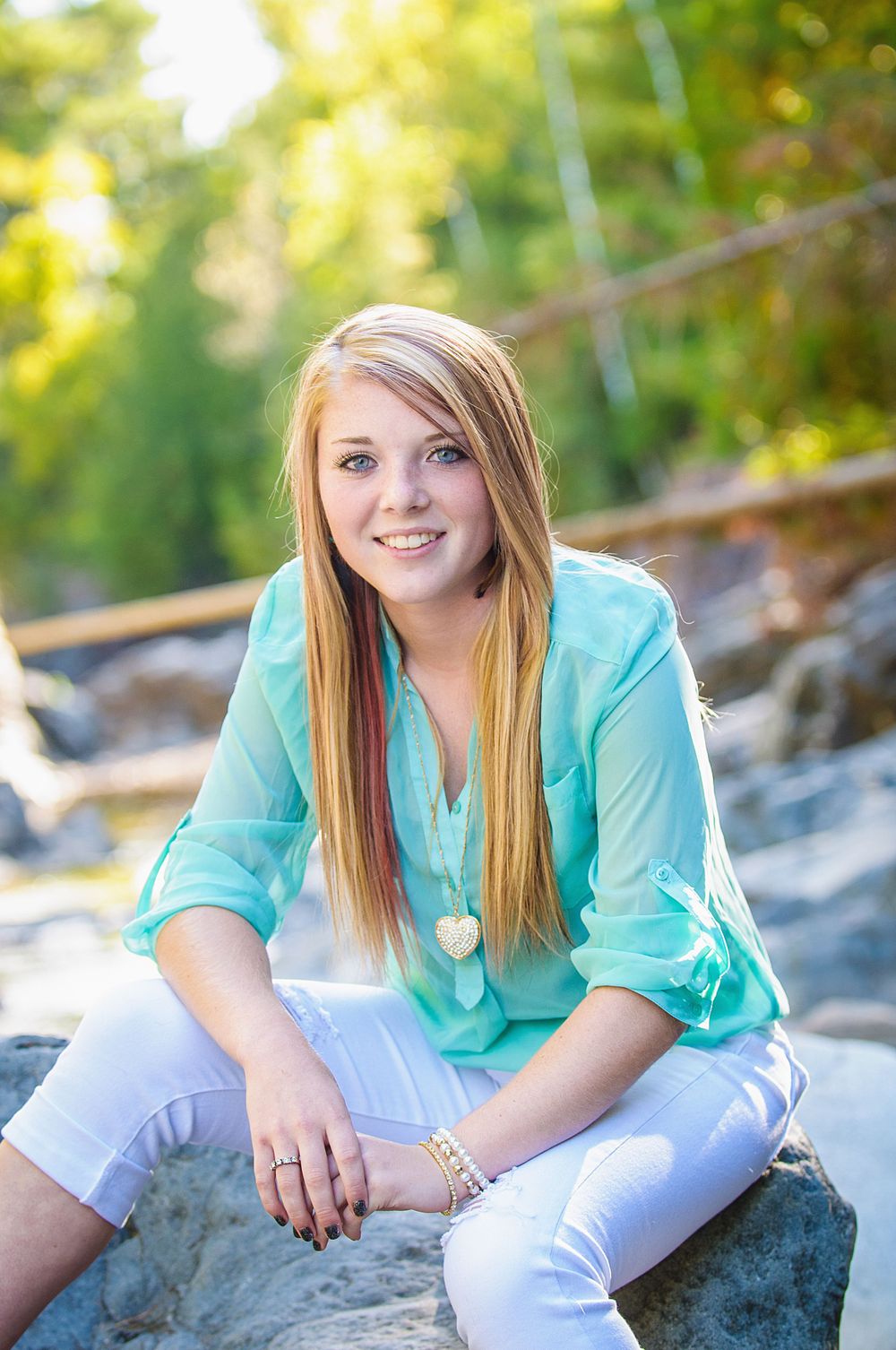 Smiling teenage girl in a teal blouse and white jeans sitting on rocks outdoors for a senior portrait session.