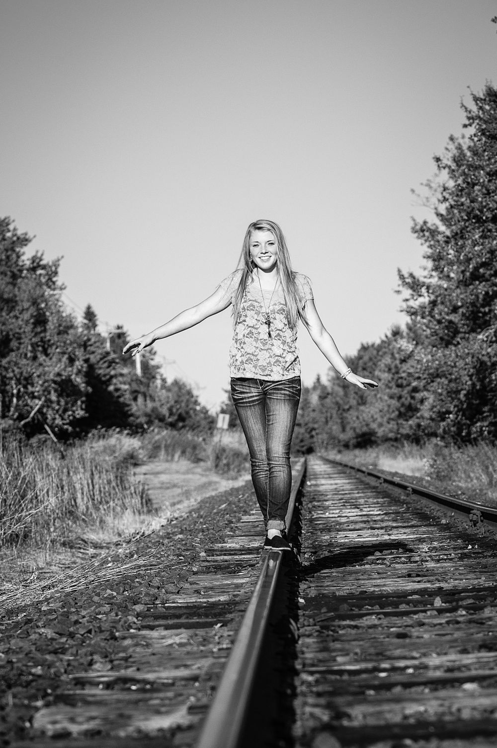 Black and white photo of a smiling blonde teen balancing on railroad tracks surrounded by trees during senior photos.