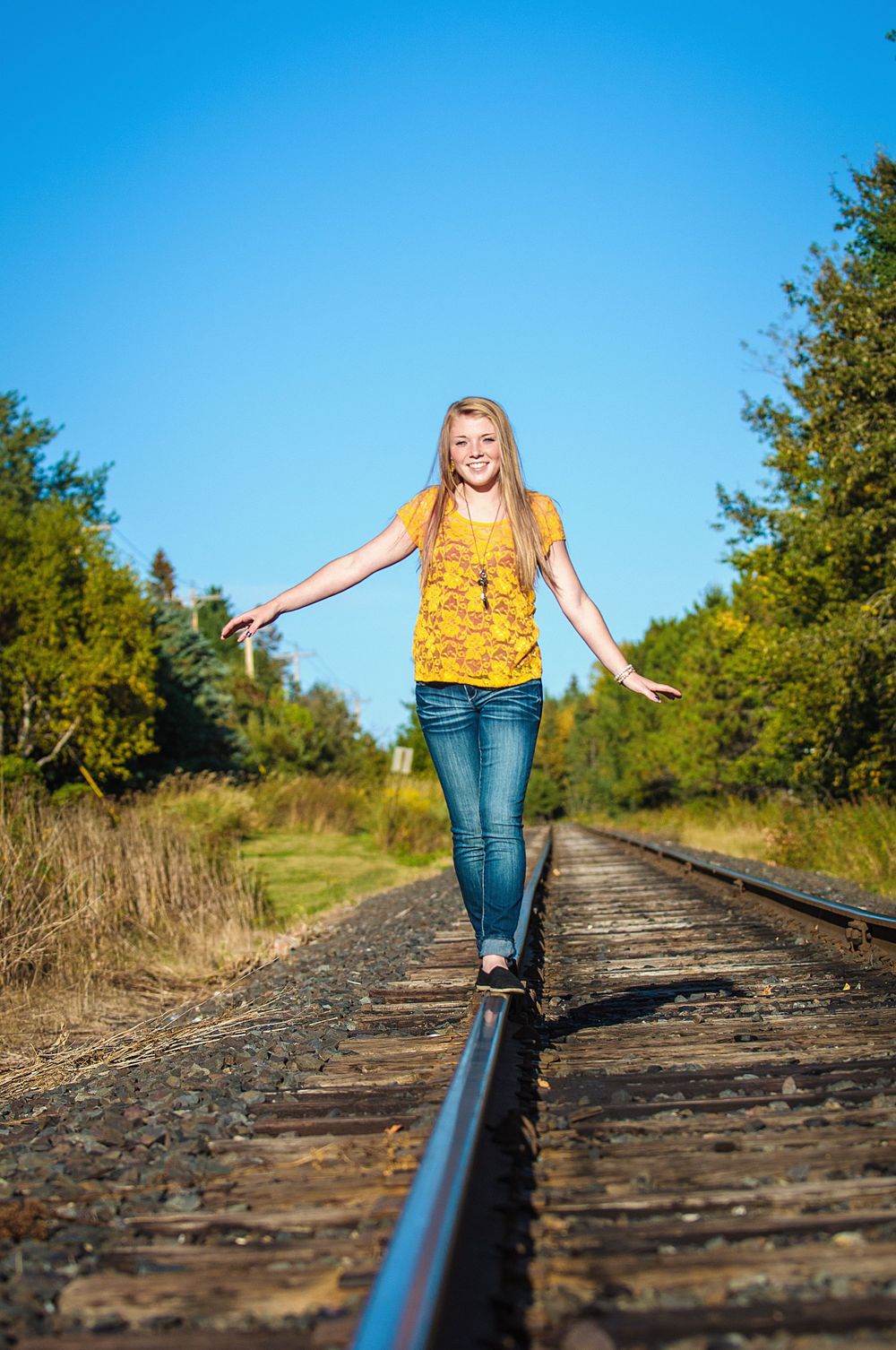 Smiling blonde teenage girl in a yellow top and jeans balancing on railroad tracks under a bright blue sky.