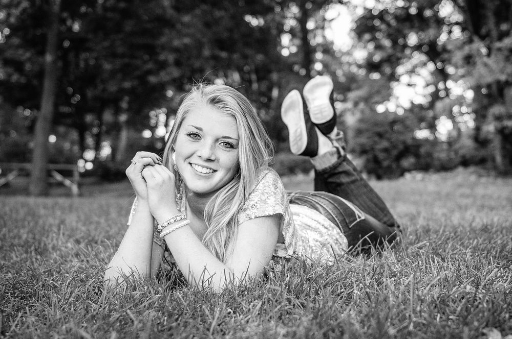 Black and white senior portrait of a smiling blonde girl lying on grass with her feet up in a park.