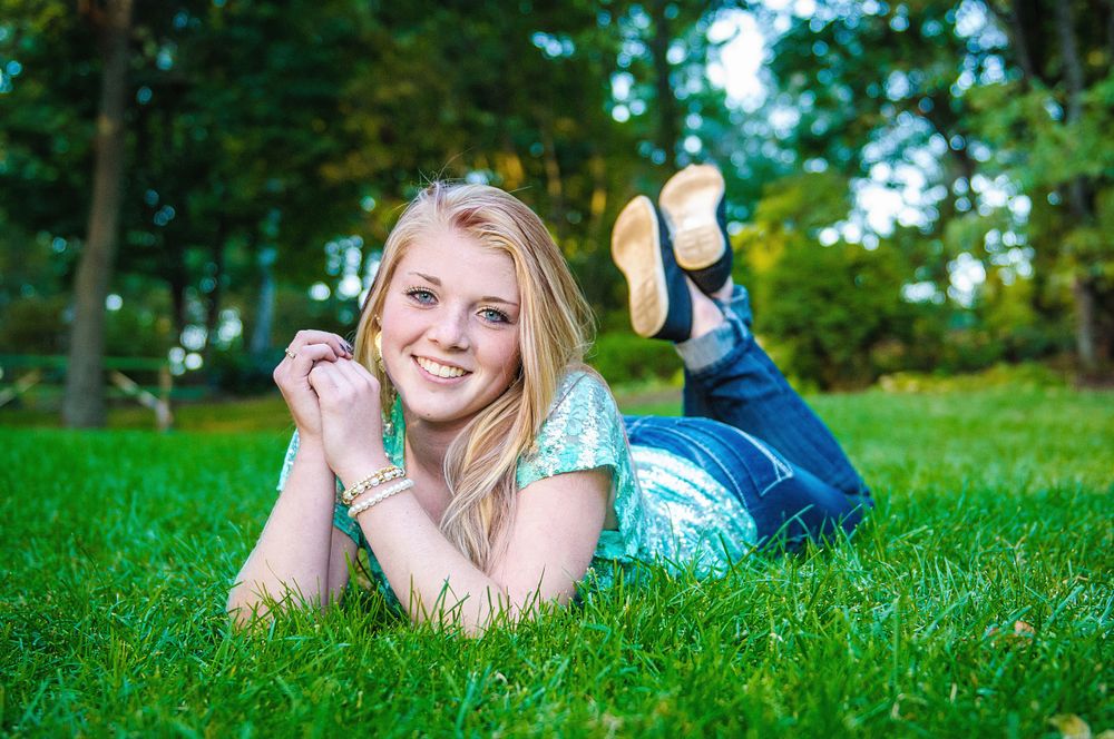 Smiling blonde teenage girl lying on green grass with feet up during an outdoor senior portrait session.