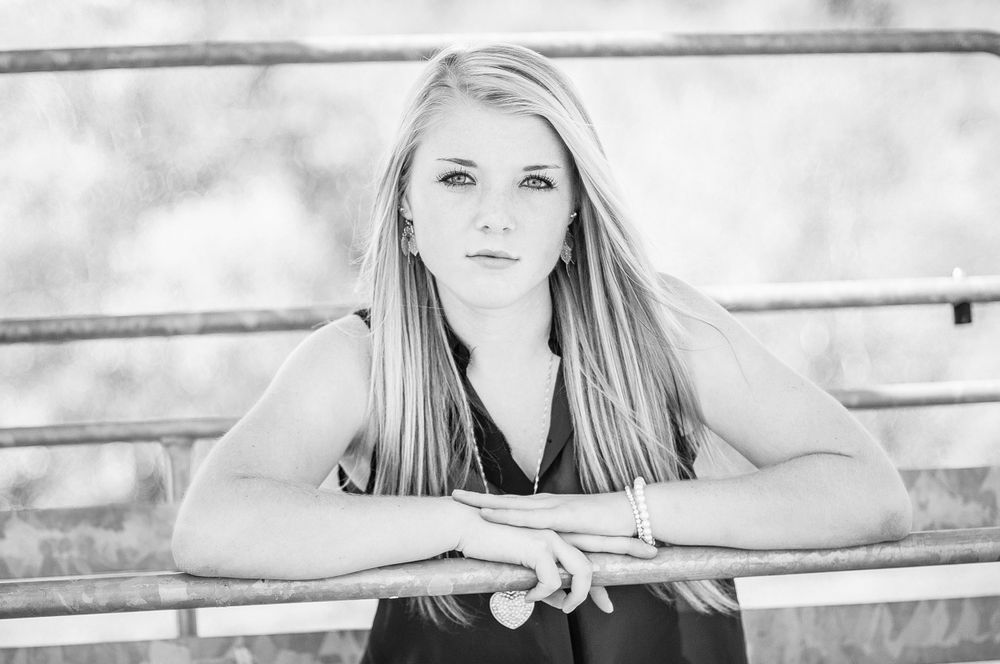 Black and white senior portrait of a serious blonde teenage girl leaning on a railing with arms crossed.