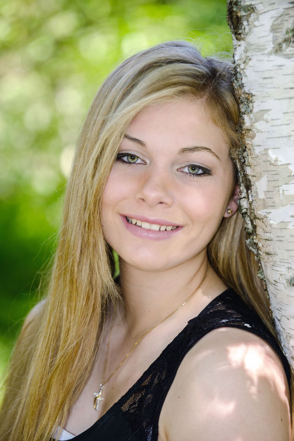 Smiling blonde teenage girl leaning against a birch tree in a lush green park during a senior portrait session.
