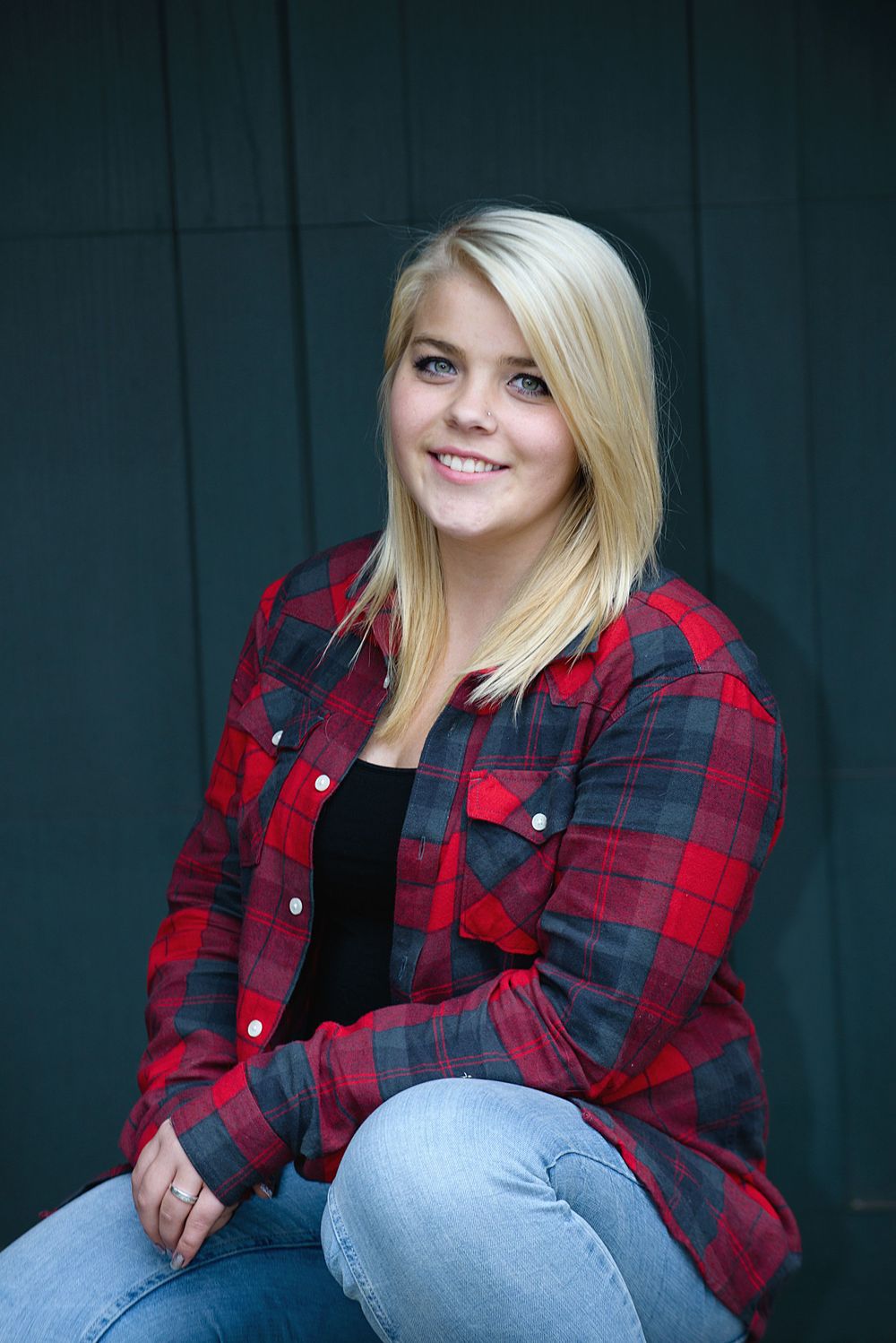 Smiling blonde teenage girl in red plaid flannel shirt sits casually against teal wooden wall.