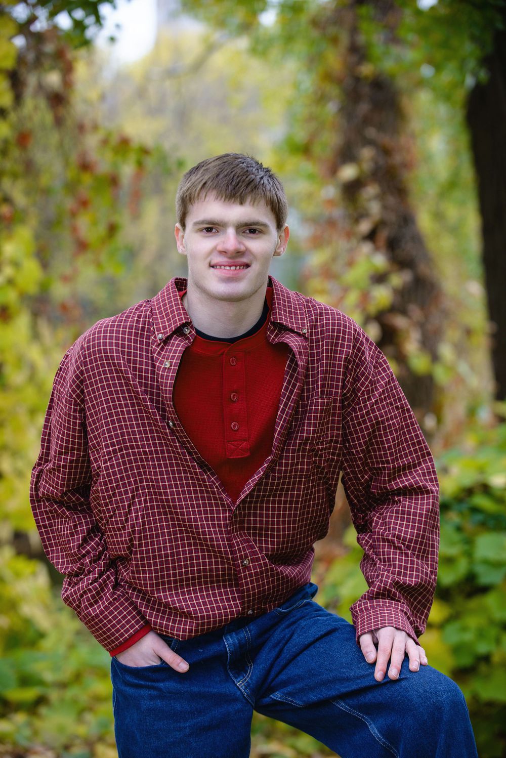 Smiling young man in red plaid shirt poses casually outdoors among autumn foliage.
