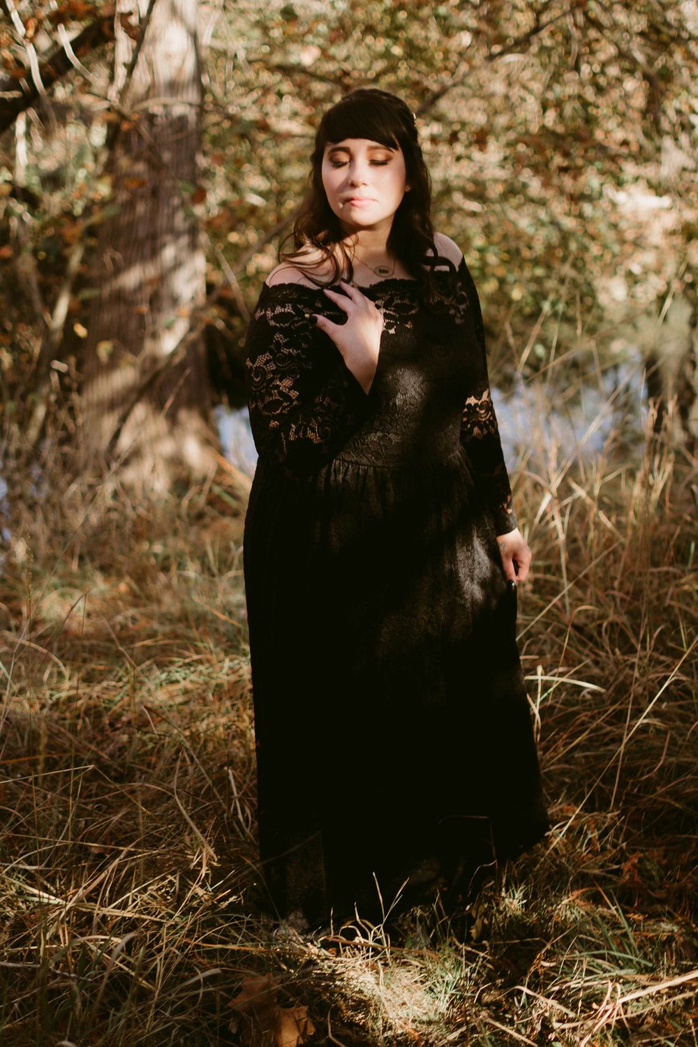 An ethereal portrait of someone in a black lace dress posed thoughtfully in dappled forest light.
