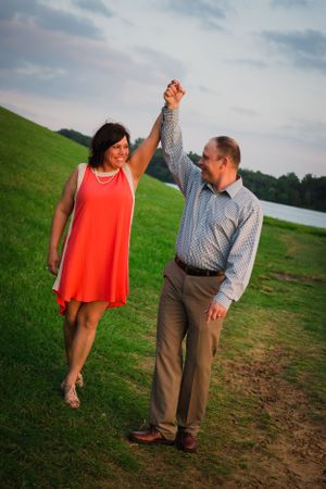 A couple dances joyfully on a grassy hillside near a lake at sunset.