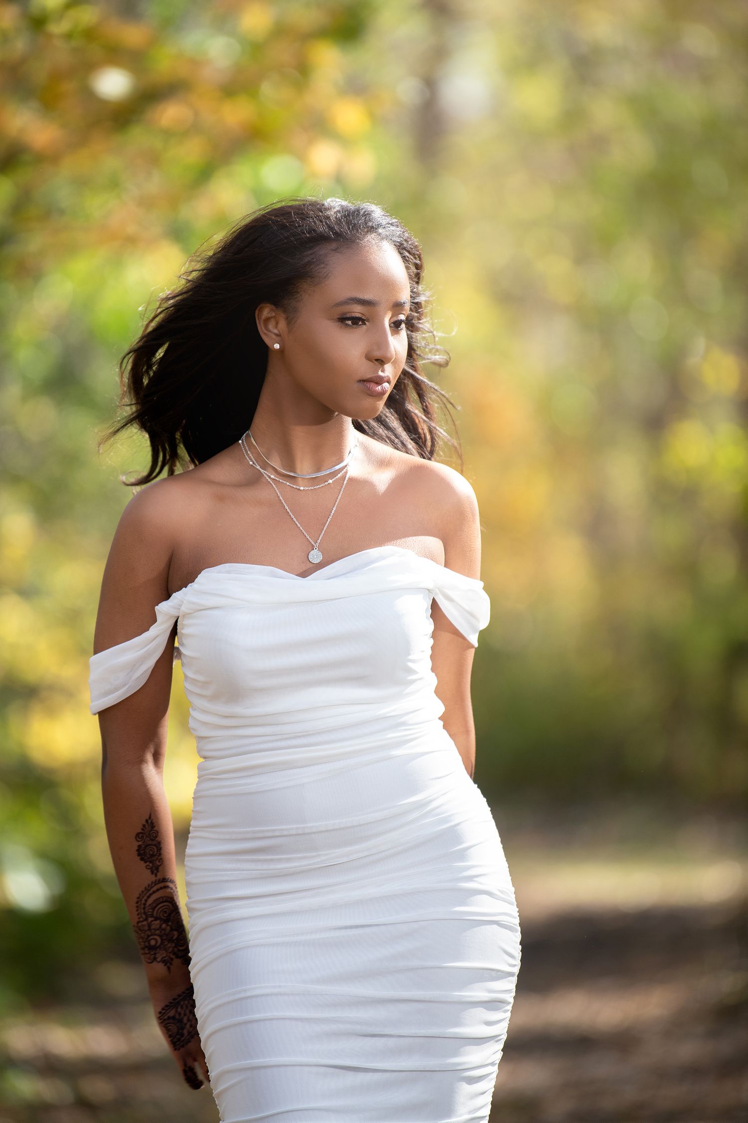Woman in elegant white off-shoulder dress poses in autumn woodland setting.