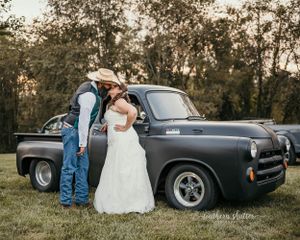 Two people stand next to a vintage gray car at sunset, dressed in western and formal attire in an outdoor setting.