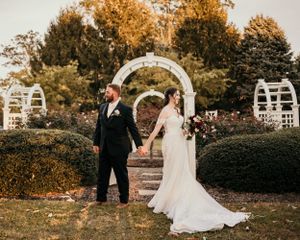 A couple holds hands during their wedding ceremony under a garden archway surrounded by autumn trees at sunset.