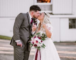 A bride in a white lace gown shares a kiss with her groom in a grey suit while holding a blush and burgundy bouquet.