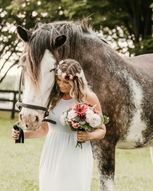 A person in a white dress holds a pink bouquet while standing beside a dappled gray horse in a sunlit field.