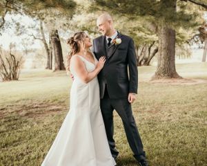 A couple in wedding attire shares a tender moment in a sunlit park setting with tall trees in the background.