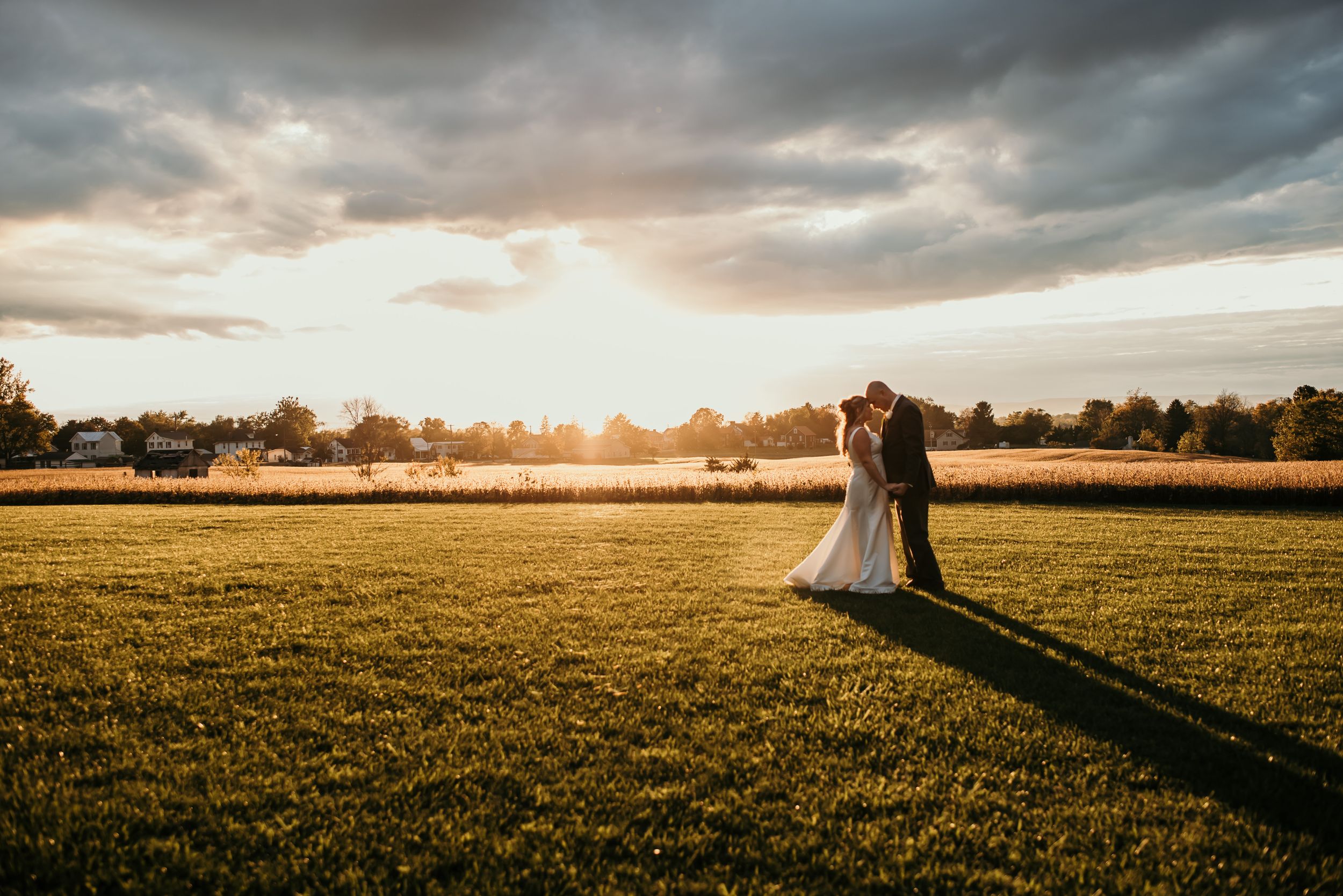 A couple embraces in a golden field at sunset with dramatic clouds and long shadows stretching across the landscape.