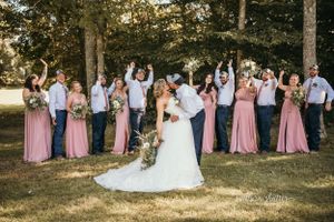 Wedding party in pink dresses and grey suits poses joyfully on sunny grass lawn during outdoor celebration.