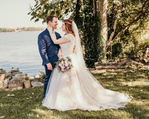 A couple poses in wedding attire by a lakeside on a sunny day, surrounded by trees and natural scenery.