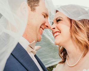 A happy wedding couple shares a joyful moment under a flowing veil as they smile at each other.
