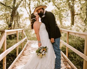 A couple shares a romantic kiss on a rustic wooden bridge while holding a wedding bouquet in a sunlit forest setting.