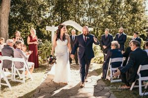 A couple walks together down an outdoor aisle lined with white chairs and wedding guests during a sunlit garden ceremony. Rockfield Manor