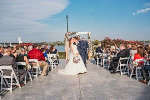 An outdoor wedding ceremony on a rooftop venue with white chairs and guests gathered under a blue sky.