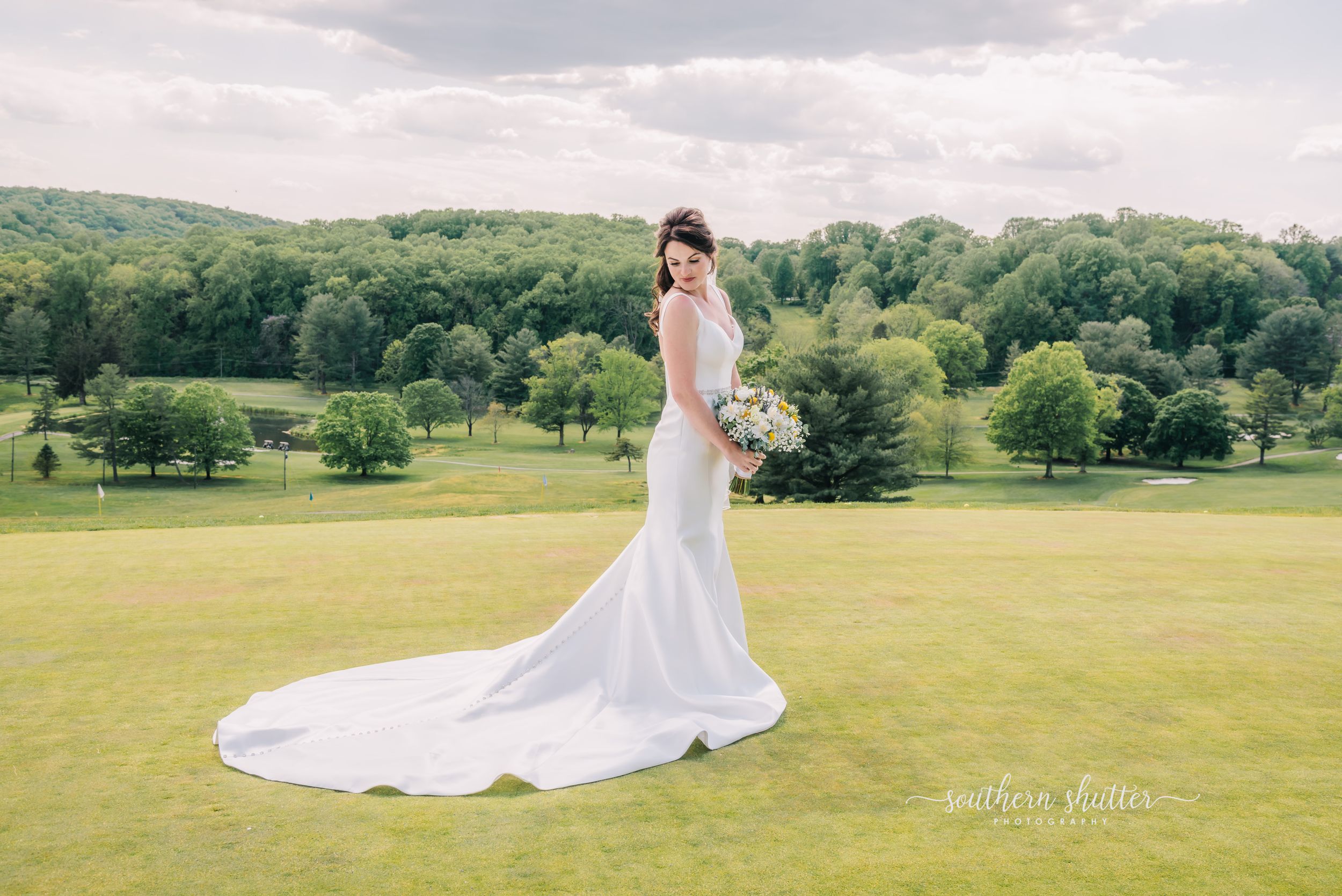 A bride in a flowing white mermaid gown stands on a lush green golf course with rolling hills and trees in the background.