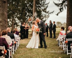 Outdoor autumn wedding ceremony surrounded by towering pine trees with guests seated in white folding chairs. Drumore Estate