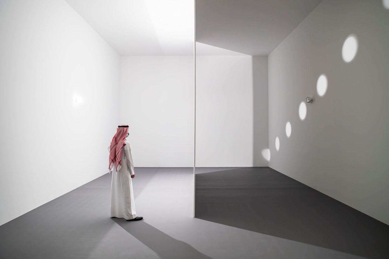 Person in traditional Middle Eastern dress views circular light patterns in a minimalist white gallery space.