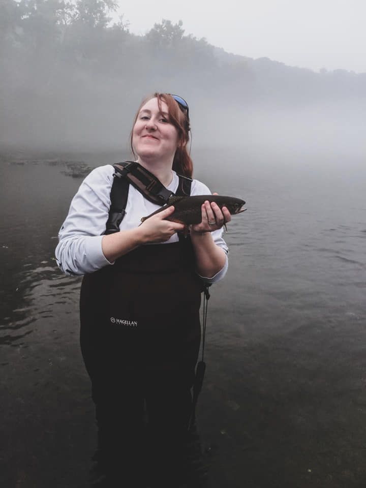 Smiling woman in waders holding a fish while standing in a misty river.