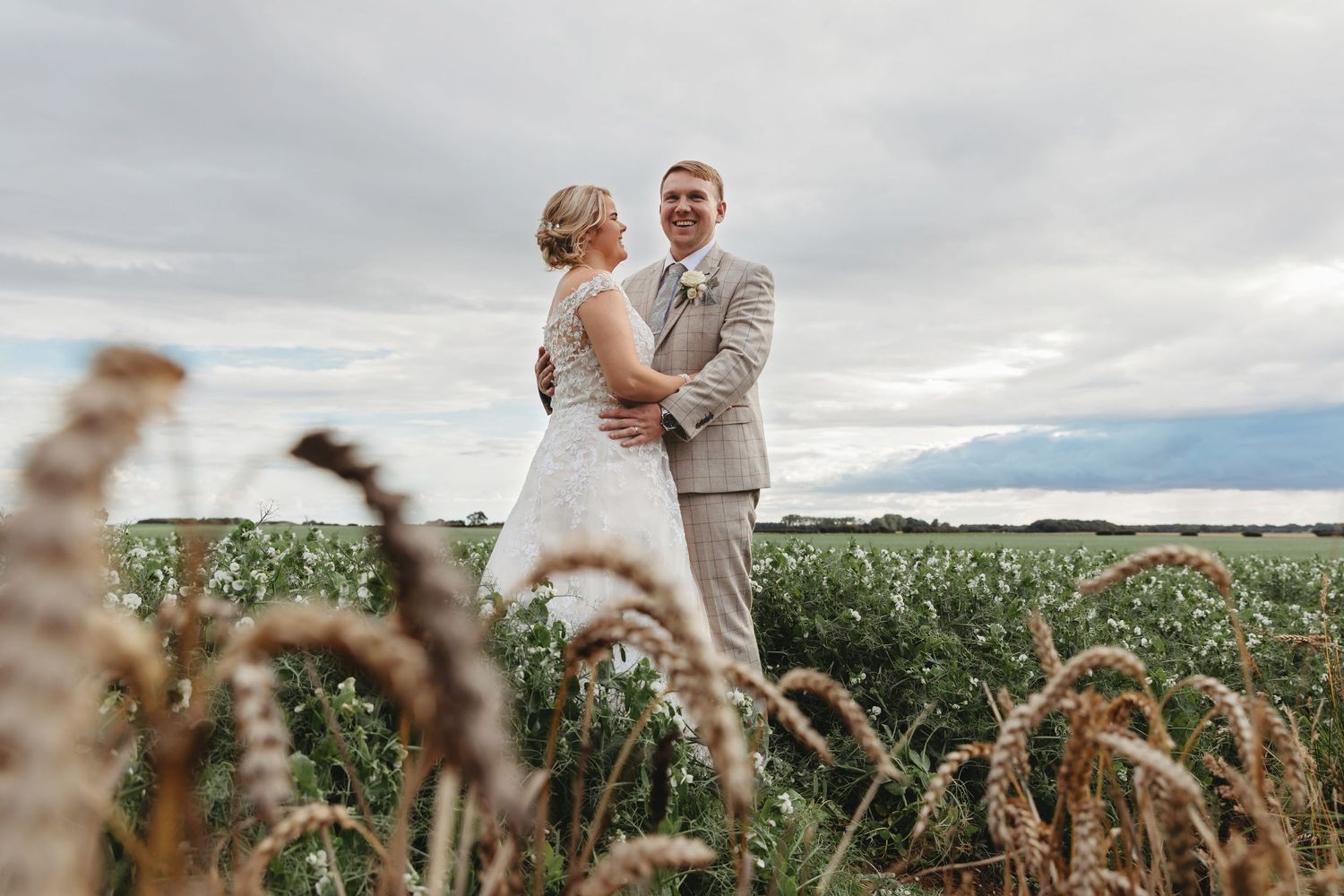A newlywed couple embraces in a field of tall grass under dramatic clouds.