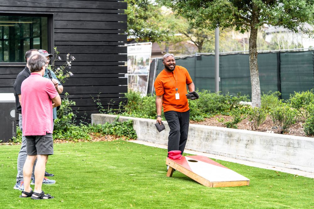Two people playing cornhole outdoors on a grassy area near a black building.