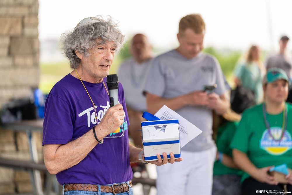 Person in purple shirt speaking into microphone while holding a blue box at outdoor gathering.