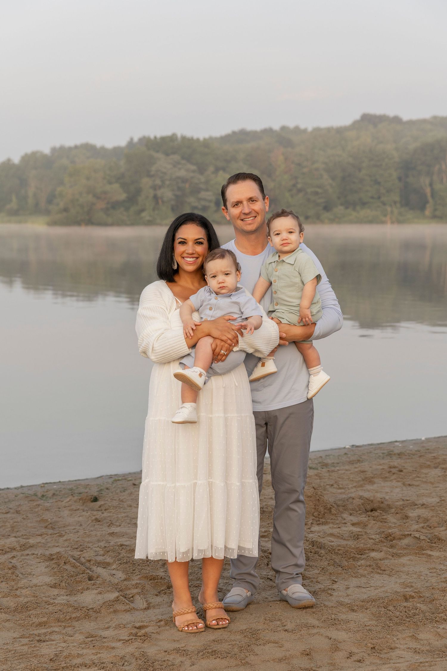 A family stands together on a beach shore at sunrise with a calm lake and wooded shoreline in the background at Stoney Creek Metropark