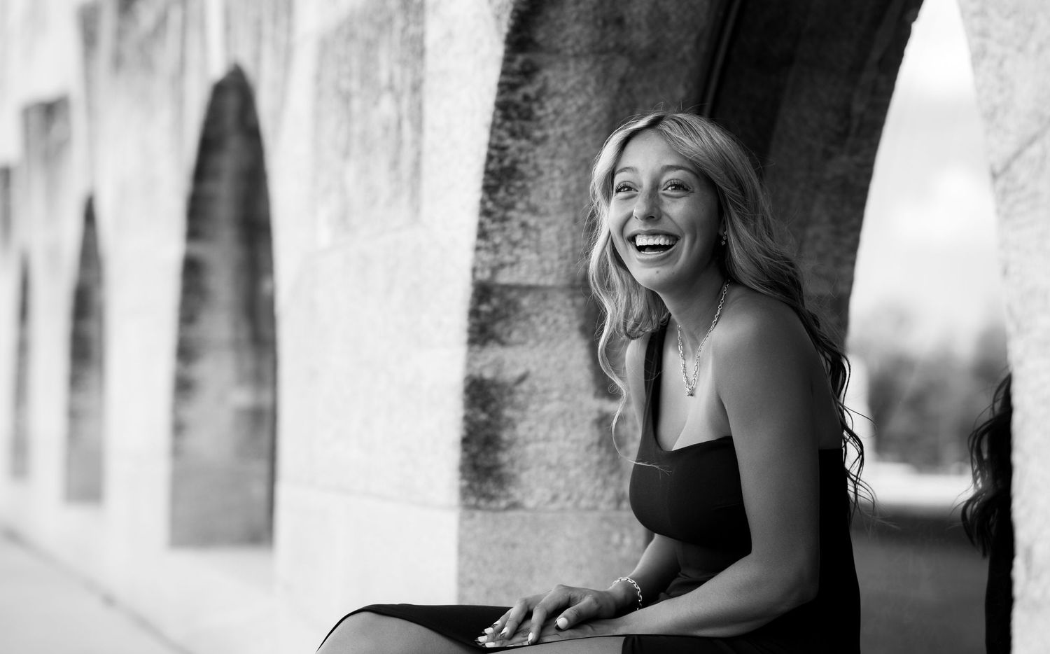 Black and white candid portrait of a person laughing while sitting outdoors near an arched wall.