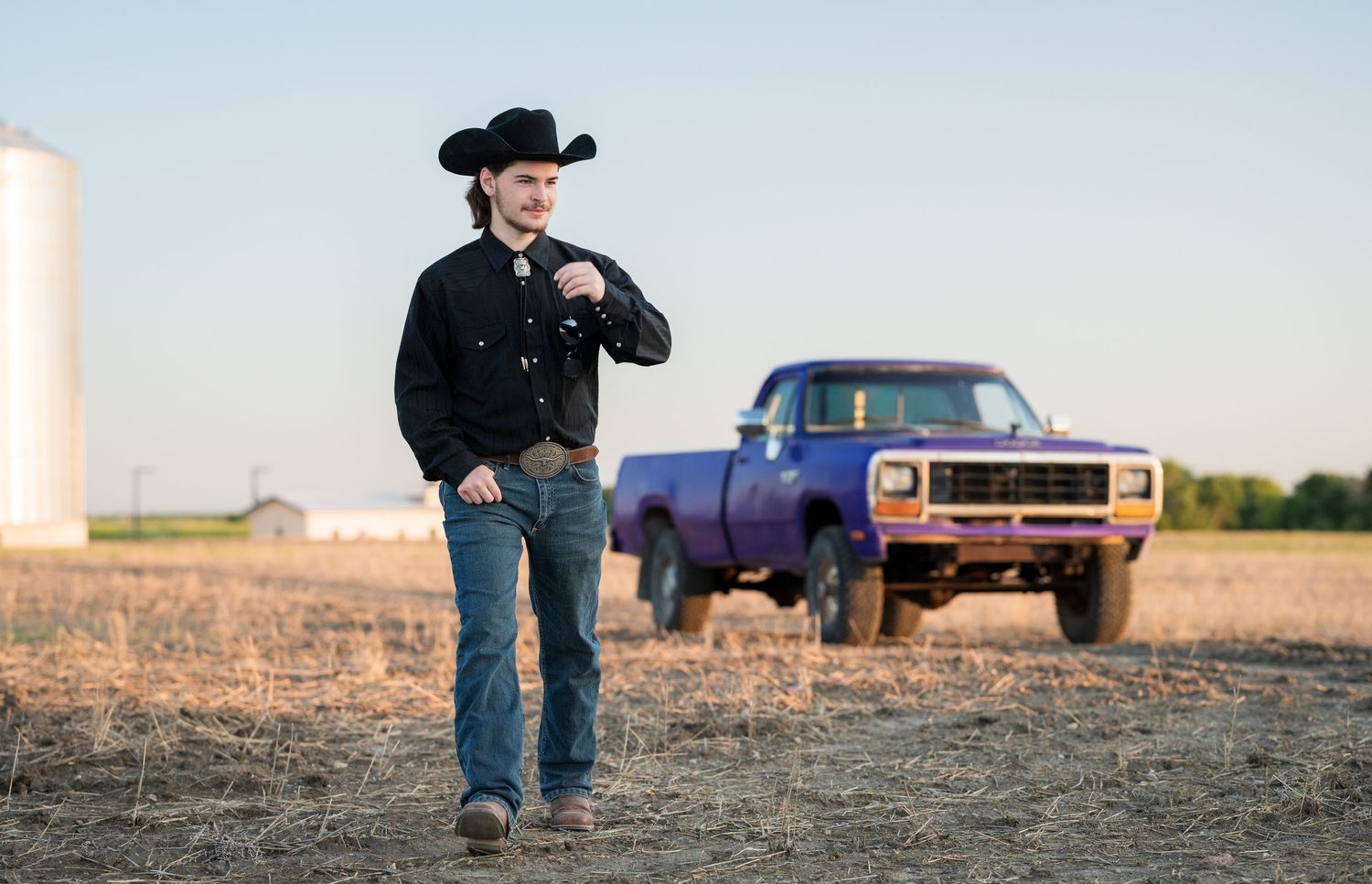 A cowboy stands in a field with a vintage pickup truck in the background during golden hour.