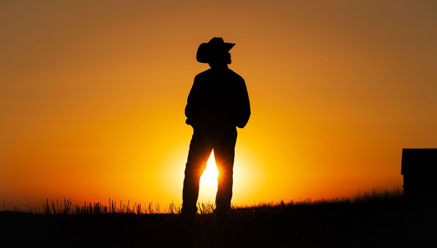 Silhouette of a person in cowboy hat standing against a vibrant orange sunset sky.