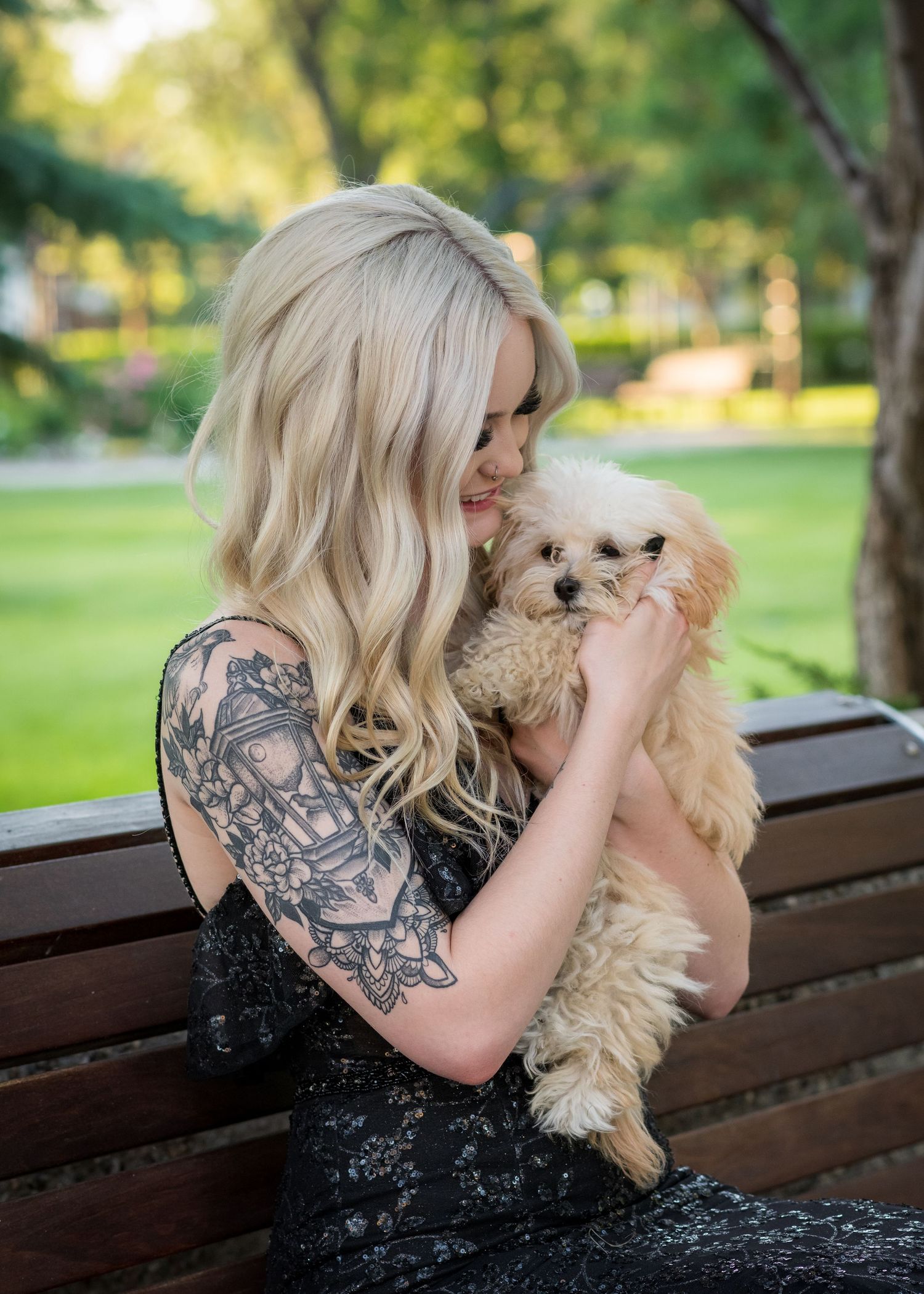 Woman in black dress holds small fluffy dog while sitting on park bench.