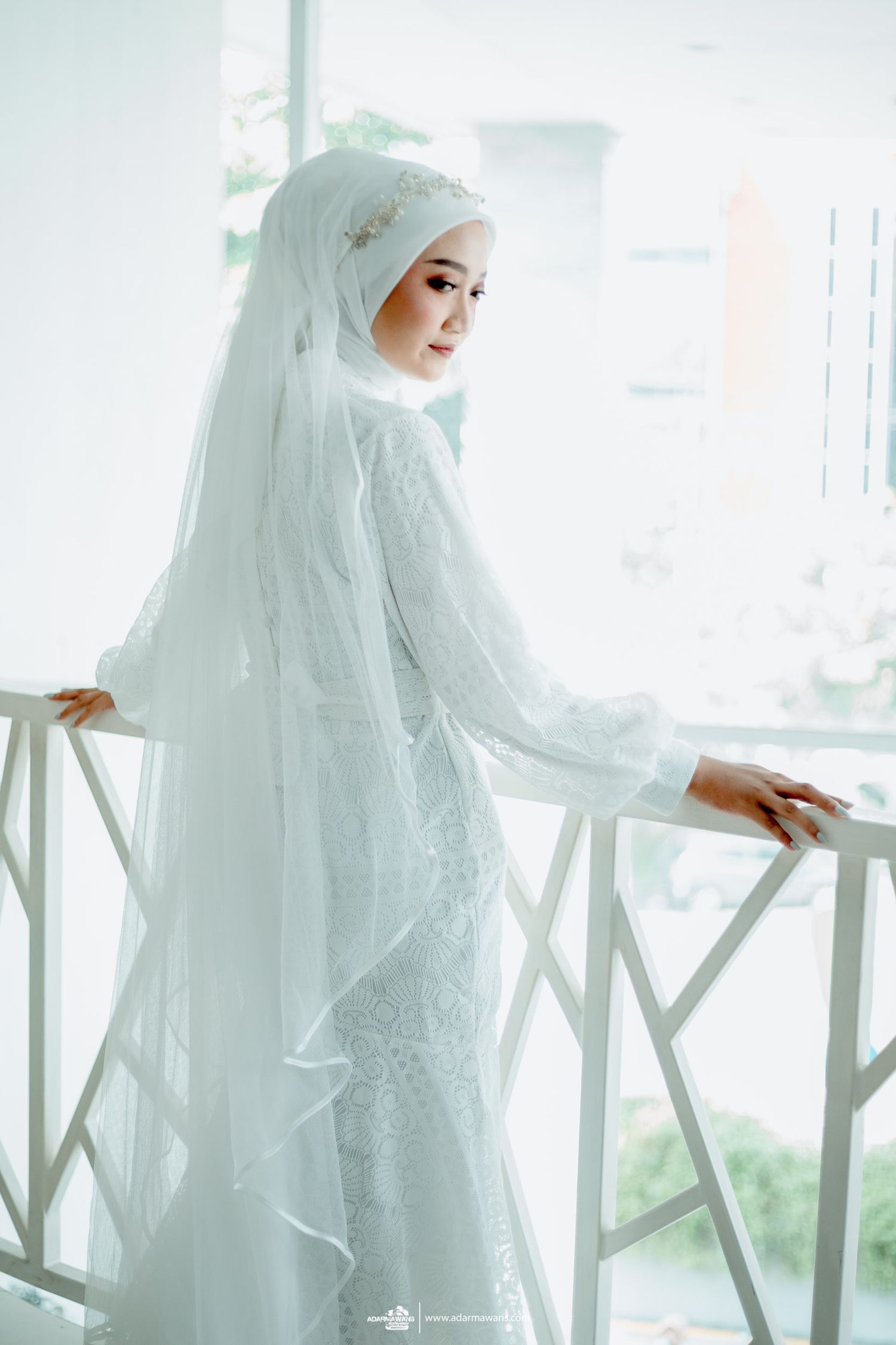Bride in detailed white lace dress and hijab poses gracefully by railing.