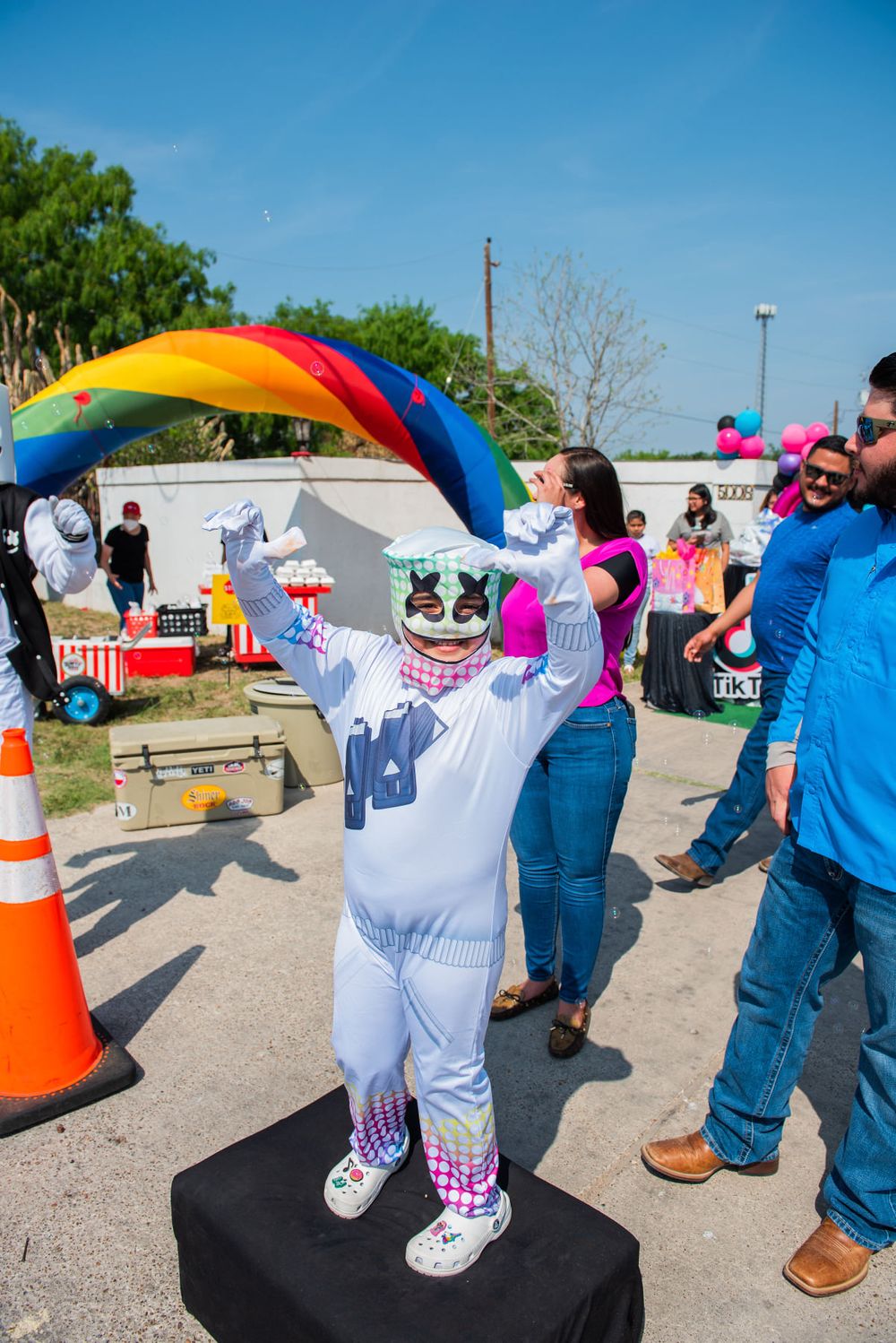 Costumed character performs under rainbow arch at outdoor event.