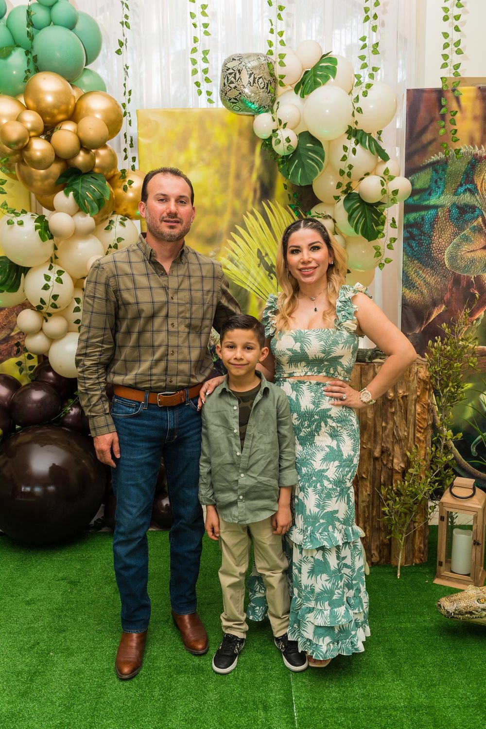 Family poses together against backdrop with gold and green balloon arch and tropical decorations.