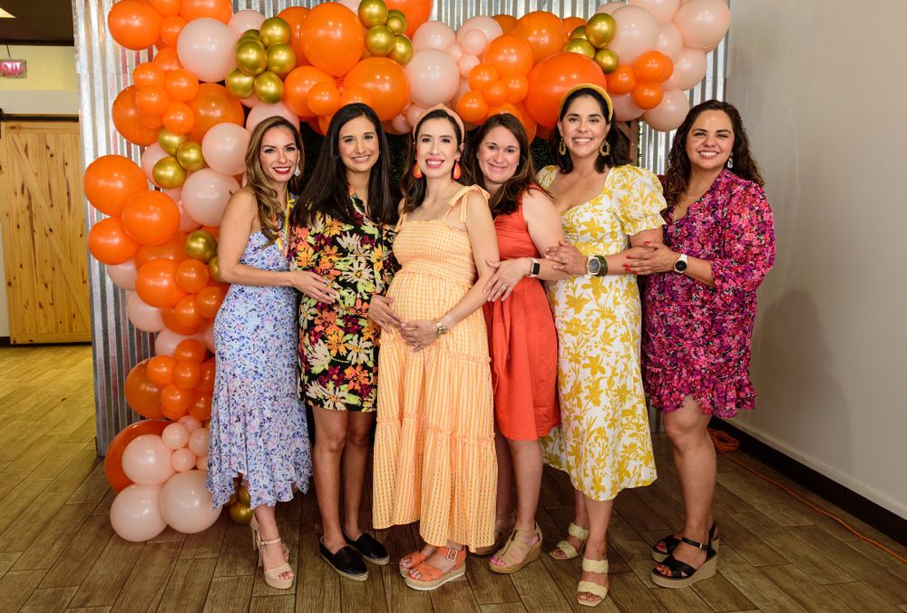 A group photo at a baby shower with orange balloon decorations in the background.