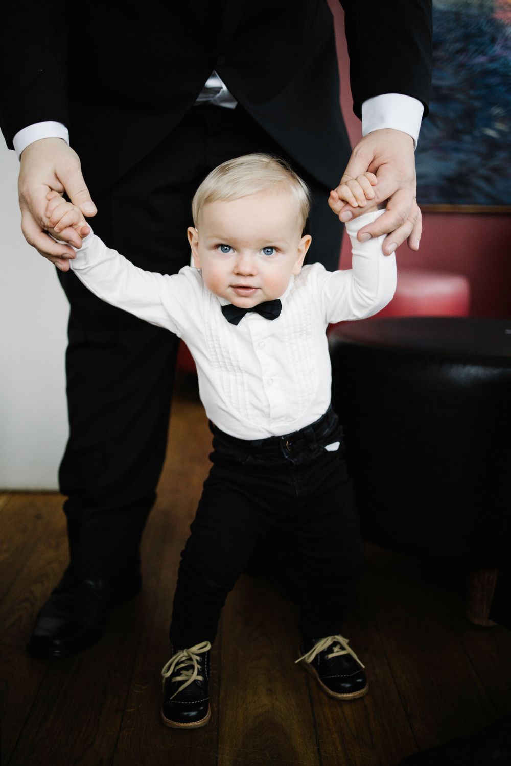A toddler in formal attire holding hands while standing on wooden flooring.