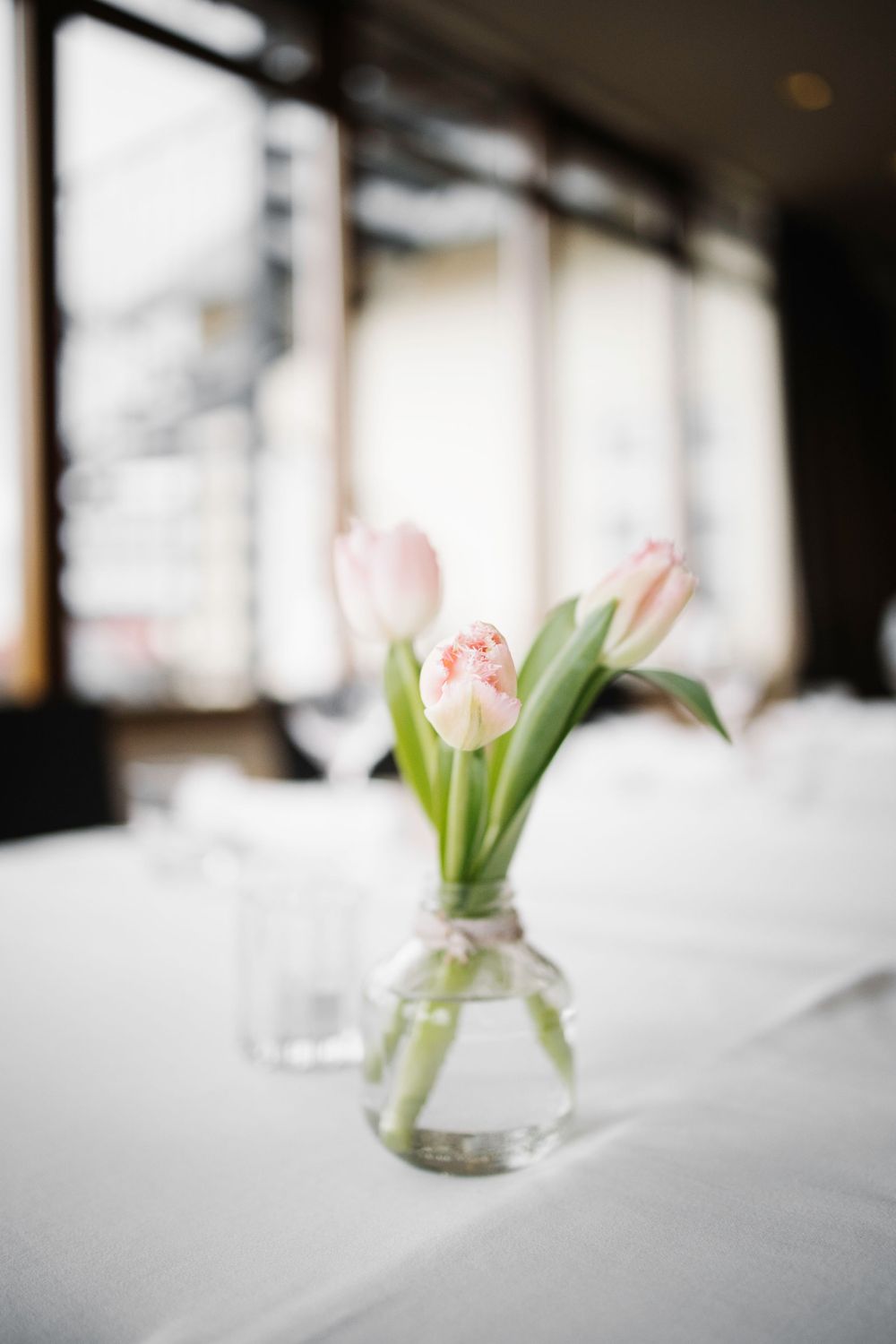 Pink tulips in a clear glass vase on white tablecloth by window.