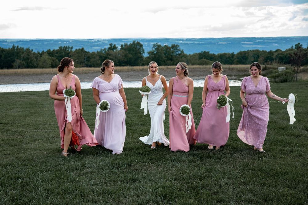 Group wearing pink dresses sits together on grassy hill during outdoor photo session.