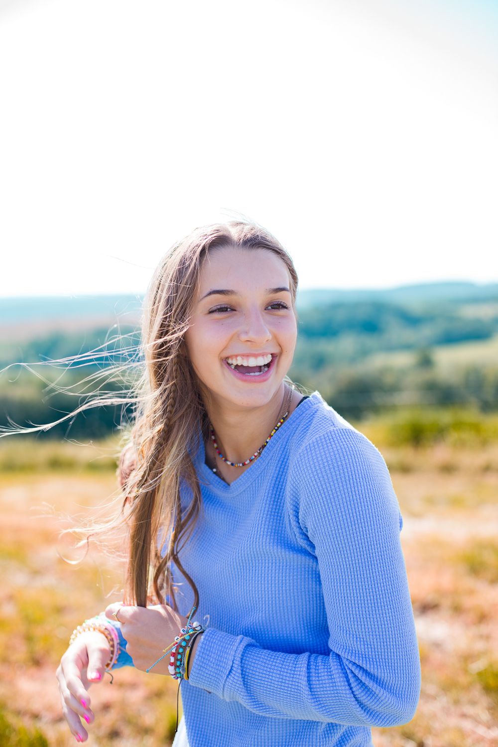 Person in blue sweater poses joyfully in an outdoor field during a sunny day photoshoot.