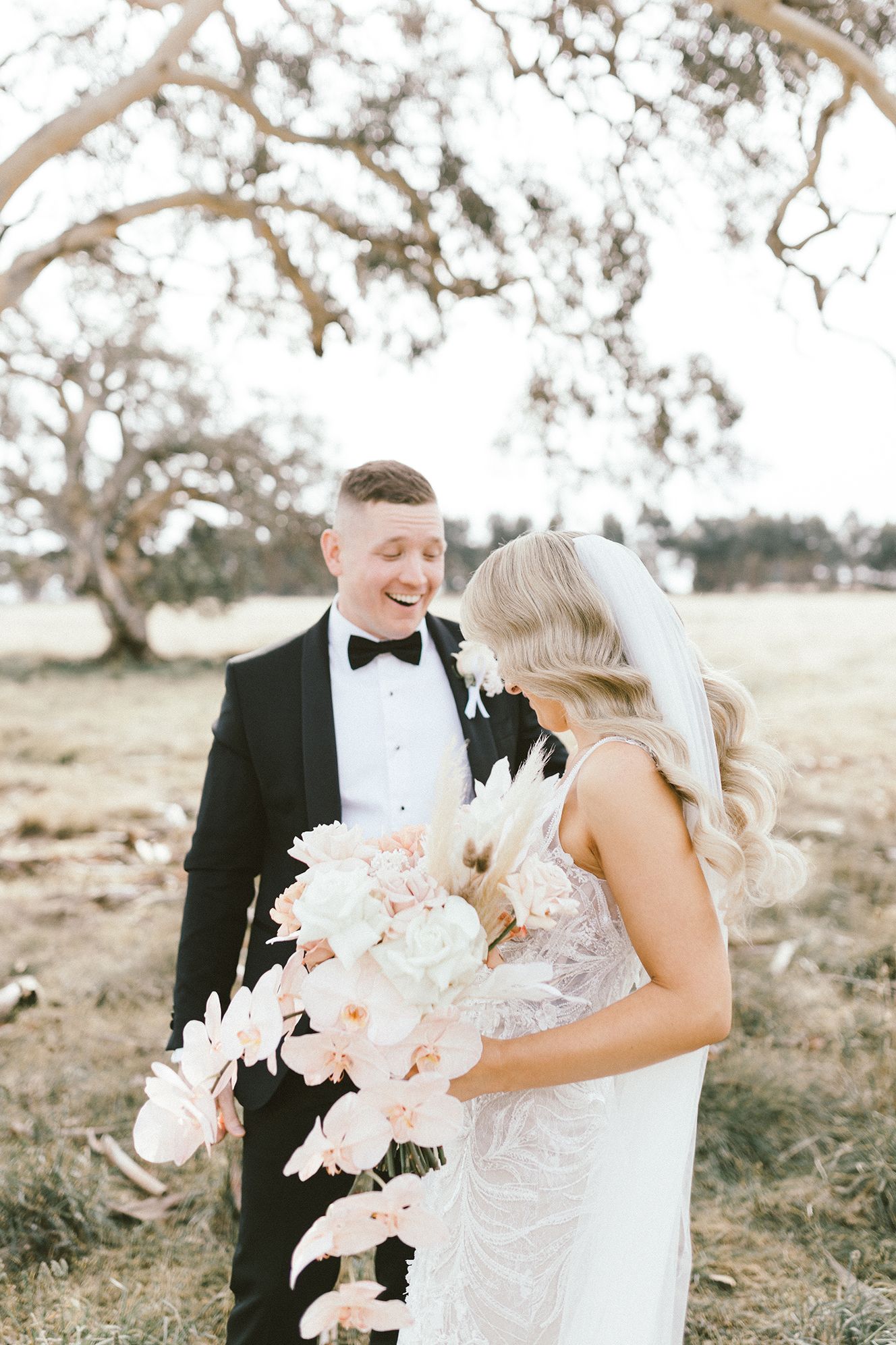 Candid moment captured during wedding photoshoot under a large tree with delicate white bouquet.