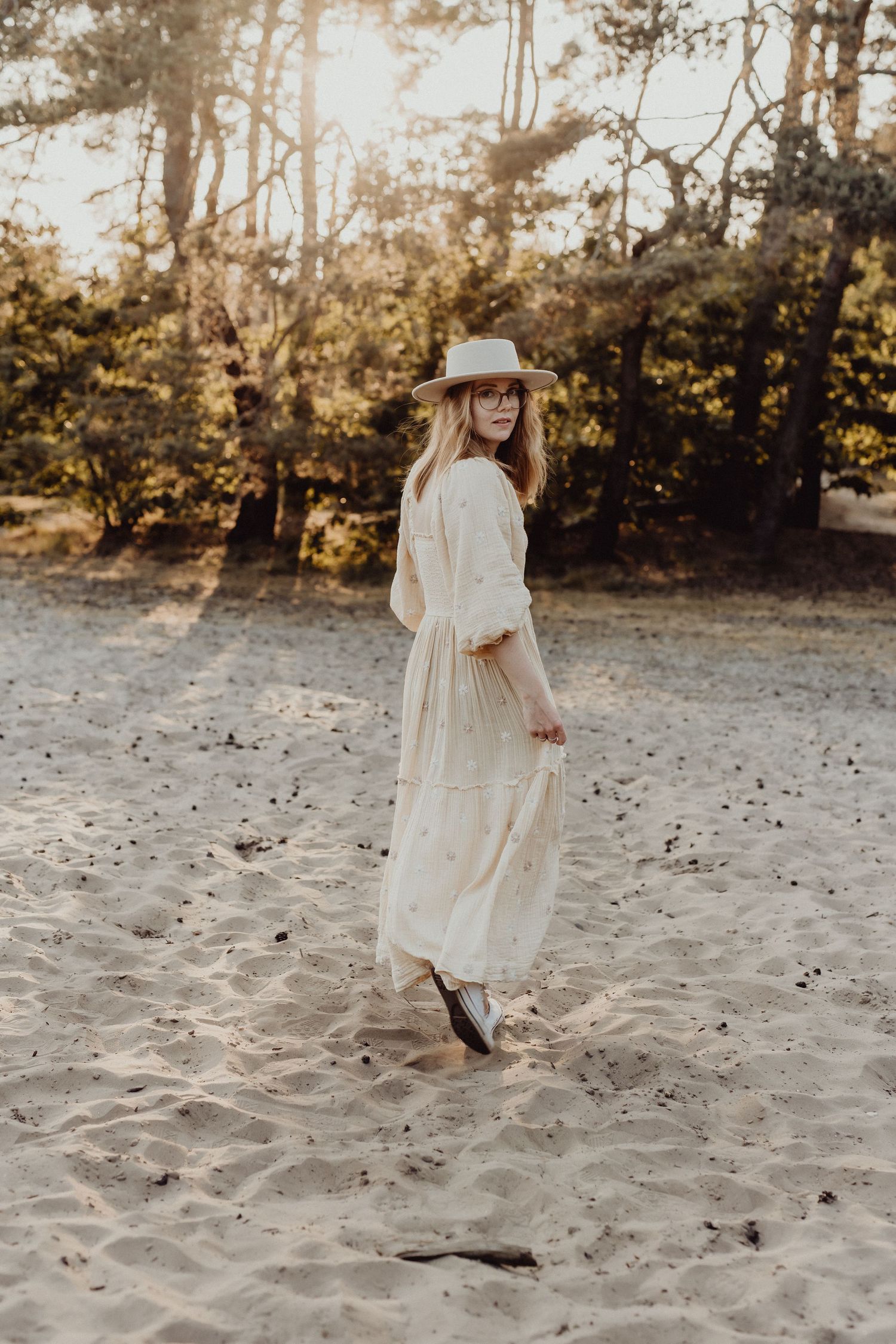 Woman in white maxi dress and hat poses on beach with trees in background.