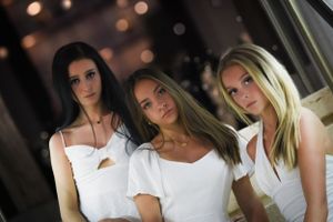 Three women in white outfits pose together with evening bokeh lights in background.