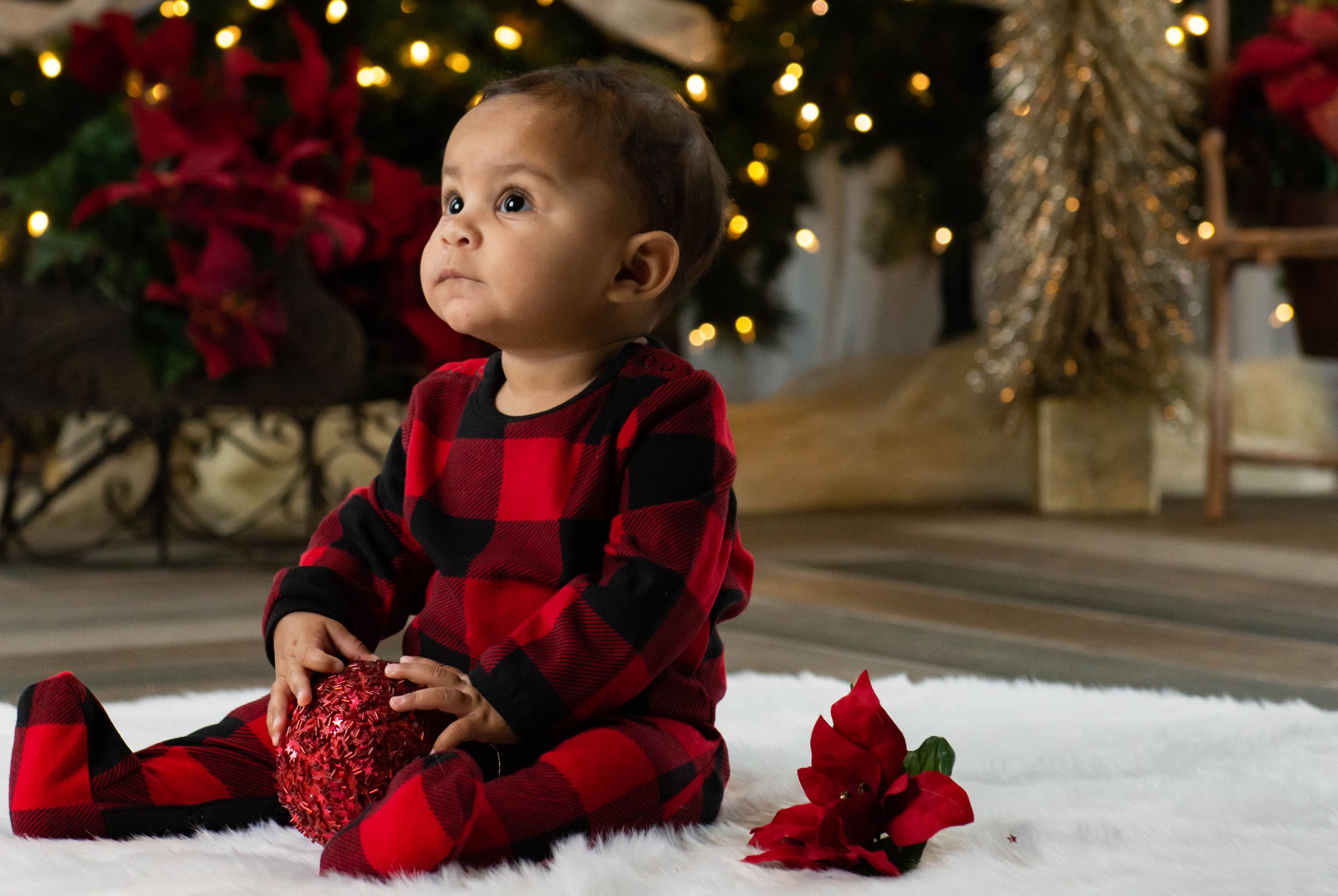 Baby in buffalo plaid pajamas sits with Christmas decorations and poinsettias. Family Photographer Batavia Cincinnati Ohio