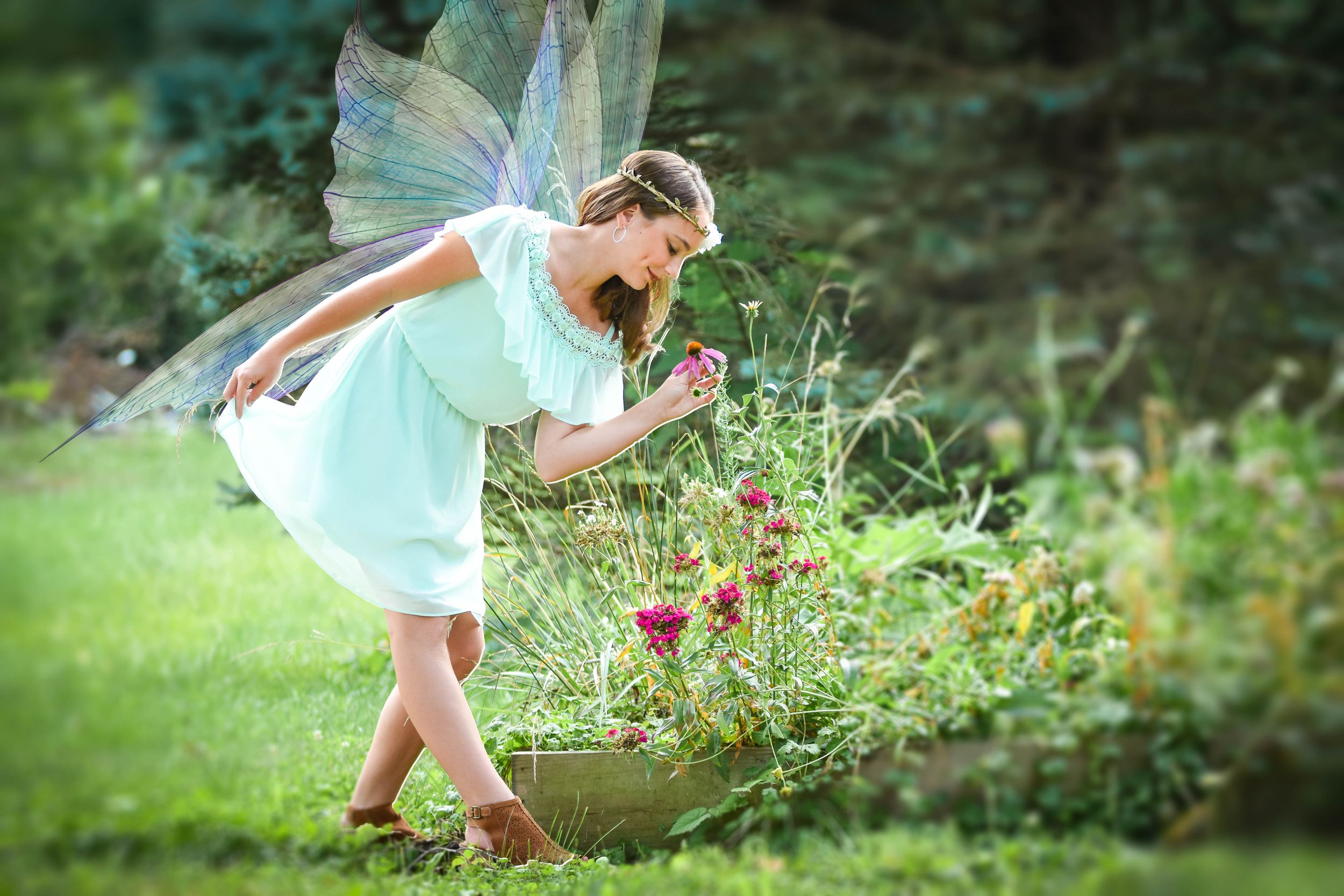 Ethereal fairy-themed portrait of someone in a mint dress with wings bending towards flowers in a garden.